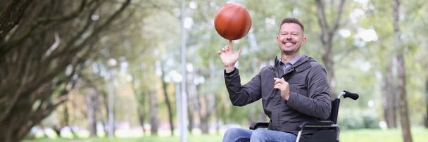 Porträt eines glücklich lächelnden Behinderten, der Basketballball wirbelt. Guy sitzt im Rollstuhl im Stadtpark. Behinderten- und Gesundheitskonzept - Foto, Bild