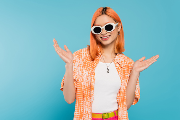 bonheur, jeune femme asiatique avec les cheveux teints debout en tenue décontractée et lunettes de soleil, geste avec les mains sur fond bleu vif, chemise orange, collier, génération z, cheveux roux  - Photo, image