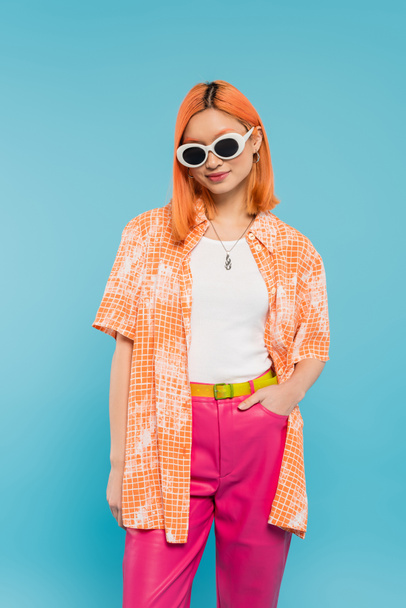 casual attire, smiling asian woman with dyed hair and sunglasses standing with hand in pocket on vibrant blue background, orange shirt, red hair, modern fashion, generation z - Фото, изображение