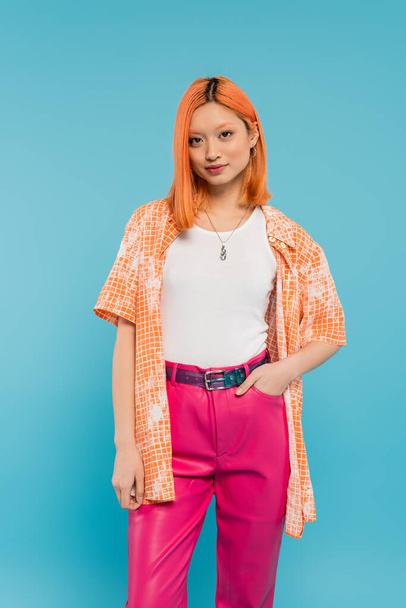 casual attire, young asian woman with dyed red hair standing with hand in pocket of pink pants on vibrant blue background, orange shirt, personal style, confidence, generation z  - Fotó, kép