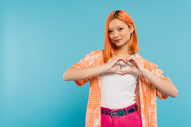 love sign, happy face, cheerful young asian woman with dyed red hair smiling and showing heart with hands on vibrant blue background, generation z, casual attire, looking at camera, young culture  - Photo, Image