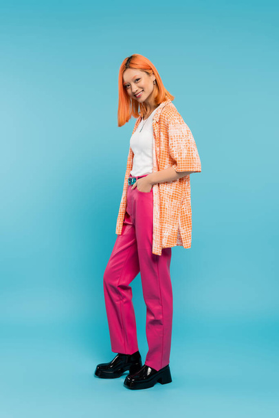 radiant smile, full length, pretty and young asian woman with dyed hair  posing with hand in pocket on blue background, looking at camera, pink pants, generation z, modern style  - Photo, Image