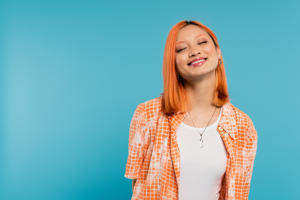 χαρούμενο πρόσωπο, λαμπερό χαμόγελο, νεαρή Ασιάτισσα με βαμμένα μαλλιά στέκεται με κλειστά μάτια σε πορτοκαλί πουκάμισο και χαμογελά σε μπλε φόντο, casual ενδυμασία, ευτυχία, ελευθερία, χαρούμενη στάση  - Φωτογραφία, εικόνα