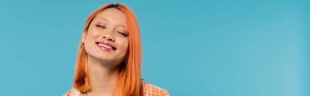 onnellinen kasvot, säteilevä hymy, nuori aasialainen nainen värjätyt hiukset seisoo suljettujen silmien oranssi paita ja hymyilee sinisellä taustalla, rento puku, onnellisuus, vapaus, iloinen asenne, banneri  - Valokuva, kuva