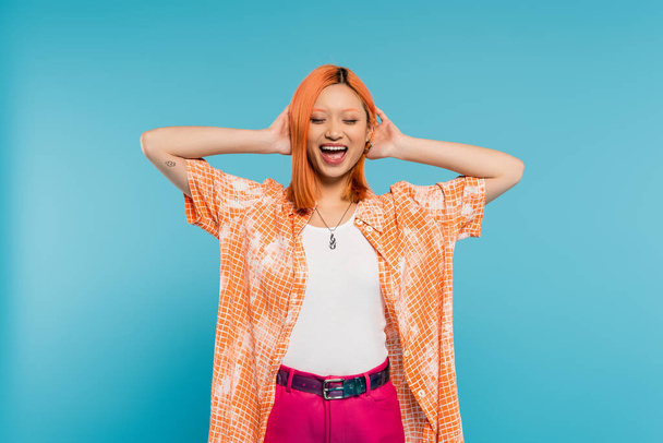 positiviteit en geluk, jonge aziatische vrouw met geverfd haar staande met gesloten ogen in oranje shirt en lachend op blauwe achtergrond, casual kleding, vrijheid, vrolijke houding, tatoeage  - Foto, afbeelding