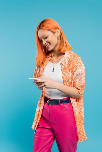 redes sociais, mulher asiática alegre com mensagens de cabelo tingido, usando smartphone, de pé sobre fundo azul, sorrindo, camisa laranja, traje casual, digital nativa, generation z  - Foto, Imagem