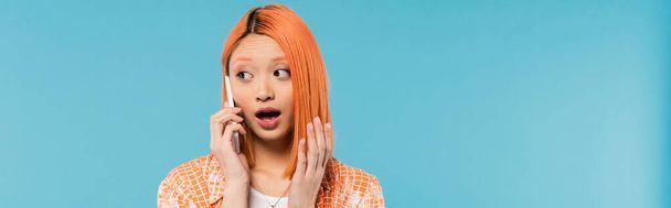 sokkolt arc, meglepett ázsiai nő telefonhívás közben, fiatal modell festett haj áll nyitott szájjal és beszél okostelefonon kék alapon, félrenéz, érzelmi, banner  - Fotó, kép