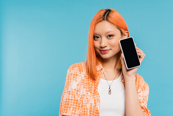 social media influencer, szczęśliwa i młoda Azjatka z farbowanymi włosami trzymająca smartfon z czystym ekranem na niebieskim tle, telefon komórkowy, kultura młodzieżowa, cyfrowy wiek, pokolenie z  - Zdjęcie, obraz