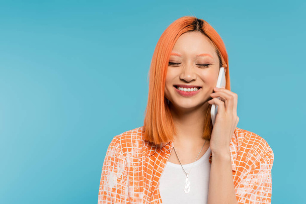 Telefonat, Positivität, fröhliche junge Asiatin mit gefärbten Haaren und geschlossenen Augen, die auf dem Smartphone auf blauem Hintergrund spricht, Mobiltelefon, Jugendkultur, digitales Zeitalter, Generation z - Foto, Bild