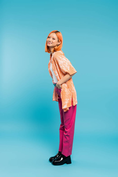 posing with hand in pocket, joyful and young asian woman with dyed hair standing in casual wear on blue background, looking at camera, pink pants, generation z, modern style, full length  - Foto, imagen