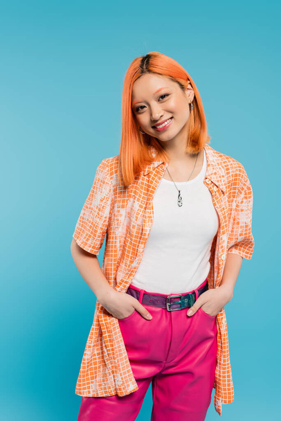 positivity, posing with hands in pockets, joyful and young asian woman with dyed hair standing in casual wear on blue background, looking at camera, pink pants, generation z, modern style  - Foto, Imagem