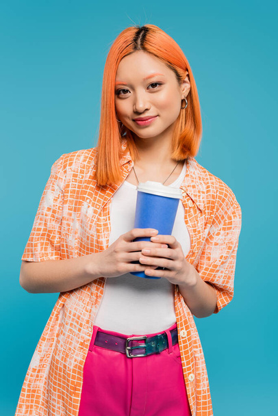 drink in hands, happy asian and young woman with red hair holding paper cup and looking at camera on blue background, casual attire, generation z, coffee culture, hot beverage, single use cup  - Φωτογραφία, εικόνα