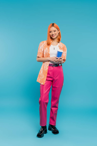 cheerful asian and young woman with red hair holding paper cup and looking at camera on blue background, casual attire, generation z, coffee culture, hot drink, single use cup, full length - Fotó, kép