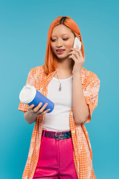 電話、赤い髪をしたアジアの若い女性が紙コップを持っていて、青い背景でスマートフォンで話している、カジュアルな服装、世代z 、コーヒー文化、熱い飲み物、シングルユースカップ  - 写真・画像