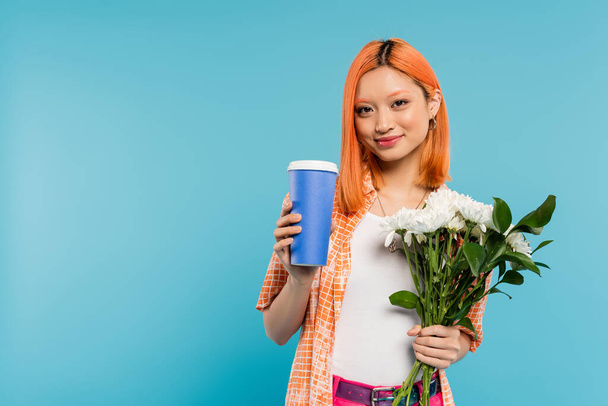 θετικότητα, ευτυχισμένη ασιατική και νεαρή γυναίκα με κόκκινα μαλλιά κρατώντας καφέ για να πάει σε χάρτινο κύπελλο και μπουκέτο λουλούδια σε μπλε φόντο, casual ενδυμασία, γενιά z, κουλτούρα του καφέ, ζεστό ρόφημα  - Φωτογραφία, εικόνα