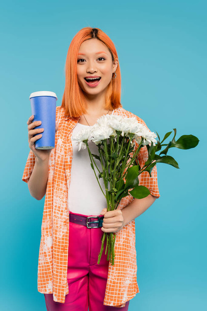 κατάπληκτος, χαρούμενο πρόσωπο, ασιατική και νεαρή γυναίκα με κόκκινα μαλλιά κρατώντας καφέ για να πάει και μπουκέτο λουλούδια σε μπλε φόντο, casual ενδυμασία, γενιά z, κουλτούρα του καφέ, ζεστό ρόφημα, χάρτινο κύπελλο - Φωτογραφία, εικόνα