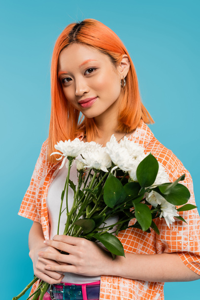 αισθησιασμός, χαρούμενη Ασιάτισσα γυναίκα με κόκκινα μαλλιά που κρατάει λευκά λουλούδια σε μπλε φόντο, casual ενδυμασία, γενιά z, floral μπουκέτο, ανοιξιάτικες δονήσεις, χαρούμενο πρόσωπο, γενιά z, νεανική κουλτούρα  - Φωτογραφία, εικόνα