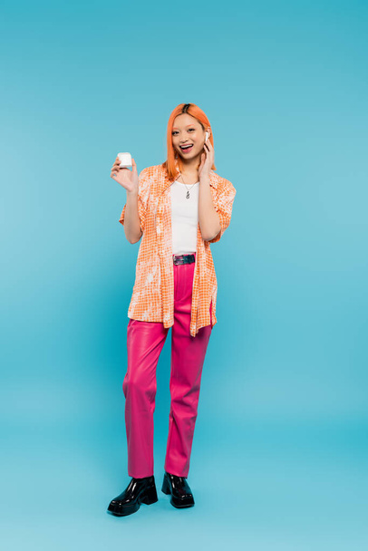 full length of happy and stylish asian woman with colored red hair, in orange shirt and pink pants holding earphone case and listening music while standing on blue background in studio - Photo, Image