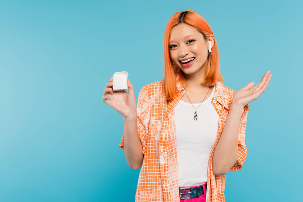 изумленная азиатка со счастливым лицом и рыжими волосами в стильной оранжевой рубашке, слушающая музыку в беспроводных наушниках, стоя на синем фоне, молодежная культура - Фото, изображение