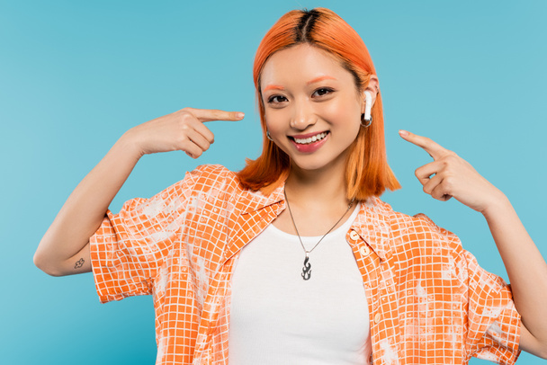 despreocupado verano, mujer asiática de moda con el pelo rojo y en blusa naranja apuntando con los dedos al auricular, escuchando música y mirando a la cámara en el fondo azul, tatuaje, generación z estilo de vida - Foto, Imagen