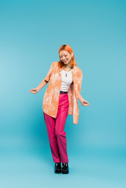 summer leisure, music lover, overjoyed asian woman in colorful clothes and wireless headphones on blue background, dyed hair, orange shirt, pink pants, gen z fashion - Foto, Imagem