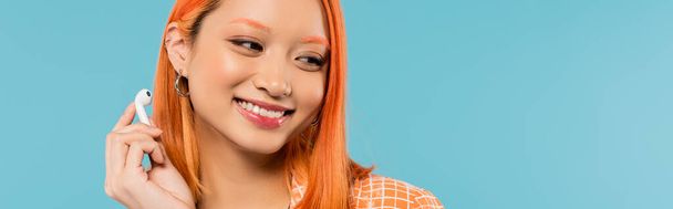 happiness and leisure, portrait of adorable asian woman with radiant smile and colored red hair holding wireless earphone on blue background, summer vibes, generation z, banner - Photo, Image