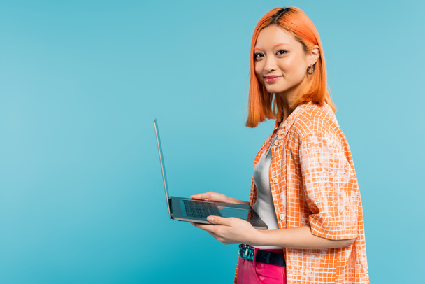 vrolijk gezicht, optimistische emotie, jeugdige aziatische vrouw met gekleurd rood haar, in oranje shirt met laptop en kijkend naar camera op blauwe achtergrond, freelance levensstijl, generatie z - Foto, afbeelding