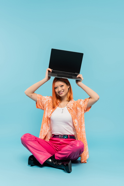 ευτυχία, ασιατική γυναίκα με χαρούμενο πρόσωπο και κόκκινα μαλλιά κρατώντας φορητό υπολογιστή με λευκή οθόνη πάνω από το κεφάλι, ενώ κάθεται με σταυρωμένα τα πόδια σε μπλε φόντο, τατουάζ, νεαρός ελεύθερος επαγγελματίας, νεανική κουλτούρα - Φωτογραφία, εικόνα