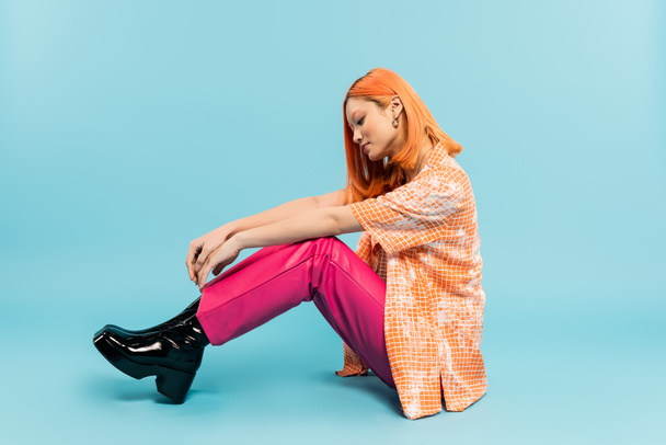 side view of young and stylish asian woman in orange shirt and pink pants sitting on blue background, dyed red hair, summer fashion, youthful style, full length, generation z - Фото, изображение