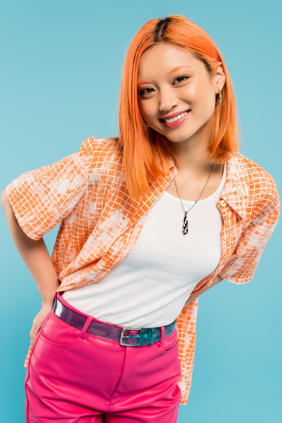 fröhliche und modische Asiatin mit gefärbten roten Haaren und strahlendem Lächeln posiert in orangefarbenem Hemd und blickt in die Kamera auf blauem Hintergrund, fröhlicher Sommer, Lebensstil der Generation Z - Foto, Bild