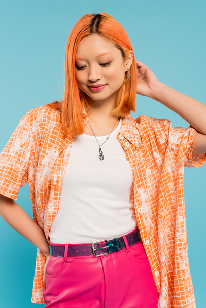 young and pleased asian woman with red colored red hair posing with hand on hip on blue background, orange shirt, pink pants, youthful fashion, summertime style, generation z - Φωτογραφία, εικόνα