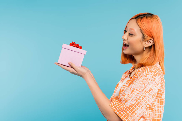 opwinding en geluk, verrukt jonge aziatische vrouw met gekleurd rood haar en open mond houden feestelijk cadeau op blauwe achtergrond, stijlvolle oranje shirt, jeugdige mode, zijaanzicht - Foto, afbeelding