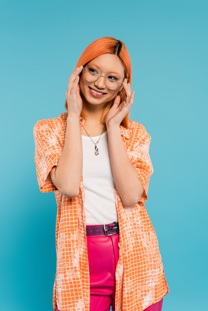 Sommermode, Glück, fröhliche Asiatin, die trendige Brillen anfasst und auf blauem Hintergrund wegsieht, gefärbte rote Haare, orangefarbenes Hemd, strahlendes Lächeln, jugendlicher Lebensstil - Foto, Bild