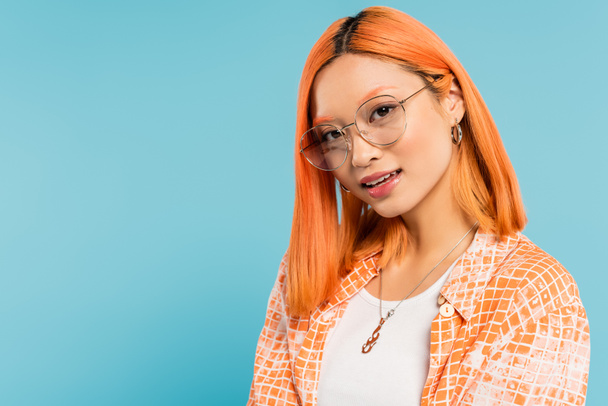 youthful fashion, happy emotion, portrait of attractive asian woman with colored red hair and expressive gaze wearing orange shirt and looking at camera on blue background, expressive gaze - Photo, Image