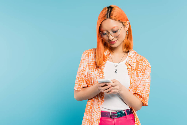 estilo de vida digital, emoción positiva, mujer asiática sonriente con el pelo rojo teñido, en gafas con estilo y red camisa naranja en el teléfono móvil sobre fondo azul, generación z, vibraciones de verano - Foto, imagen