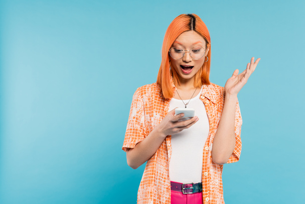 amazement, young asian woman with colored red hair and open mouth looking at mobile phone and gesturing on blue background, trendy eyeglasses, orange shirt, youthful fashion, digital lifestyle - Photo, Image
