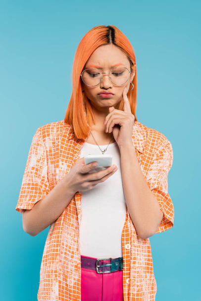 negative emotion, bad mood, displeased asian woman touching cheek while looking at smartphone on blue background, trendy eyeglasses, red colored hair, orange shirt, youthful fashion - Photo, Image