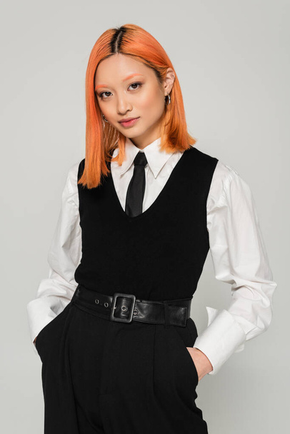 portrait of charming and young asian woman holding hands in pockets and looking at camera on grey background, dyed red hair, white shirt, black tie, vest and pants, business fashion photography - Foto, afbeelding