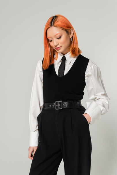 stylish and alluring asian woman with dyed red hair, in white shirt, black tie, vest and pants posing with hand in pocket on grey background, generation z lifestyle, business casual fashion - 写真・画像