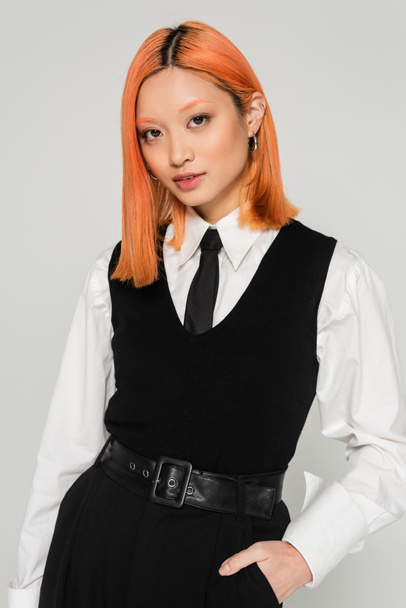 beautiful and trendy asian woman with dyed red hair, in white shirt, black tie, vest and pants holding hand in pocket and looking at camera on grey background, business casual, youth culture - 写真・画像