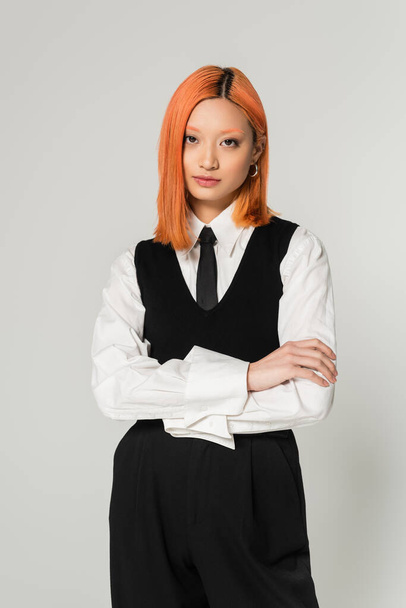 portrait of attractive and young asian woman in white shirt, black tie, vest and pants standing with folded arms on grey background, dyed red hair, confident gaze, looking at camera, business casual - Фото, зображення