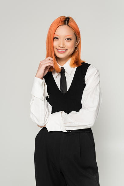 happy emotion, young and joyful asian woman with dyed red hair looking at camera on grey background, white shirt, black tie, vest and pants, business casual fashion photography, modern lifestyle - Foto, Imagem