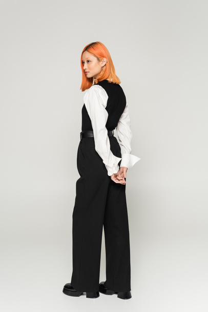 fashion photography of young asian woman with colored red hair, in white shirt, black vest and pants standing with hands behind back and looking away on grey background, business casual style - Photo, Image