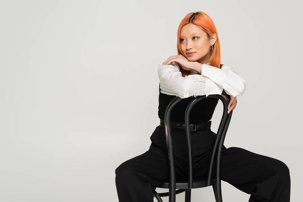 νεαρή Ασιάτισσα γυναίκα με βαμμένα κόκκινα μαλλιά κάθεται στην καρέκλα σε μαύρο και άσπρο επιχειρήσεων casual ρούχα και κοιτάζοντας μακριά σε γκρι φόντο, σύγχρονη μόδα, τη γενιά z τρόπο ζωής - Φωτογραφία, εικόνα