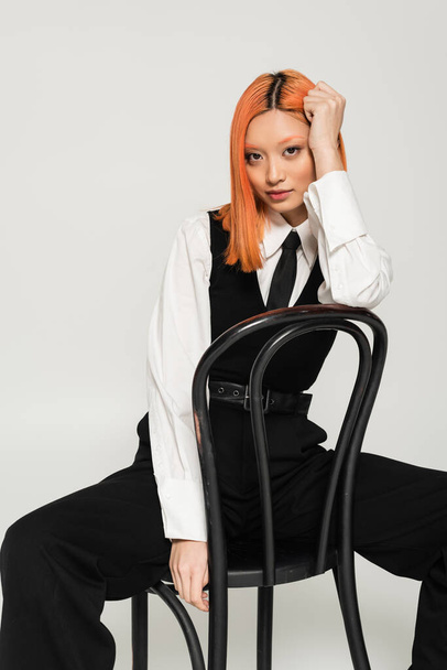 alluring and stylish asian woman with dyed red hair, in white shirt, black tie, vest and pants looking at camera while sitting on chair on grey background, generation z lifestyle, business fashion - Photo, Image