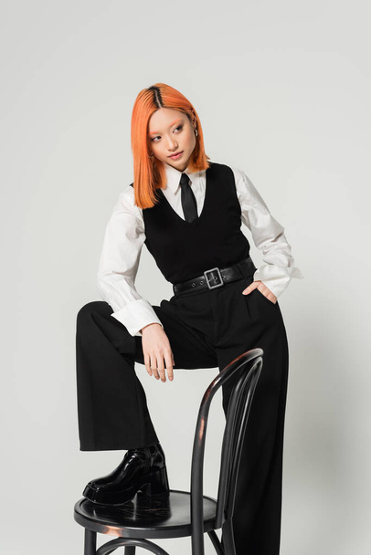 young and confident asian woman holding hand in pocket and looking away while posing with chair on grey background, dyed red hair, black and white business casual clothes, fashion shoot - Photo, Image