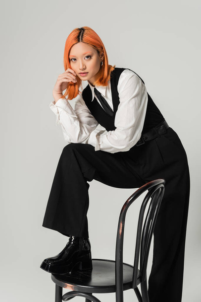 modern asian woman in fashionable black and white clothes, with colored red hair posing with chair and looking at camera on grey background, business casual fashion, youthful style - Fotoğraf, Görsel