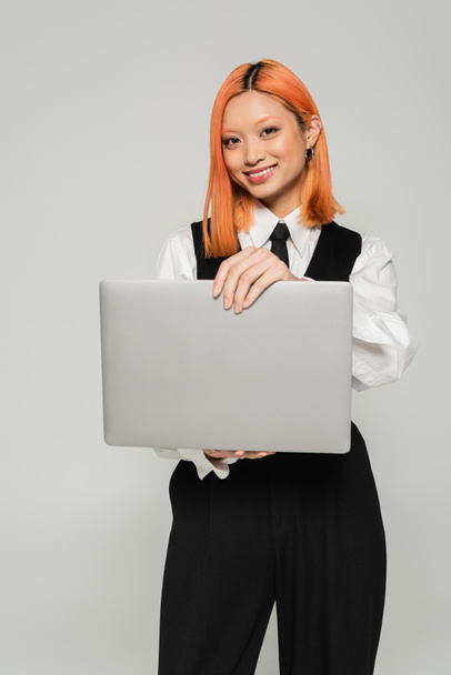 счастье, внештатный образ жизни, привлекательная азиатская женщина в черно-белой одежде держа ноутбук и улыбаясь в камеру на сером фоне, цветные рыжие волосы, деловые случайные, поколение z - Фото, изображение
