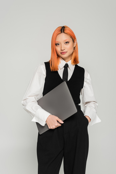 attractive asian woman with colored red hair, in black and white clothes holding laptop and looking at camera on grey background, white shirt, black vest, freelance lifestyle, generation z - Photo, Image