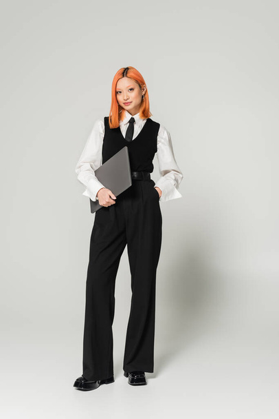 positive and confident freelancer, young asian woman with red colored hair standing with hand in pocket and laptop on grey background, business casual, white shirt, black vest and pants - 写真・画像
